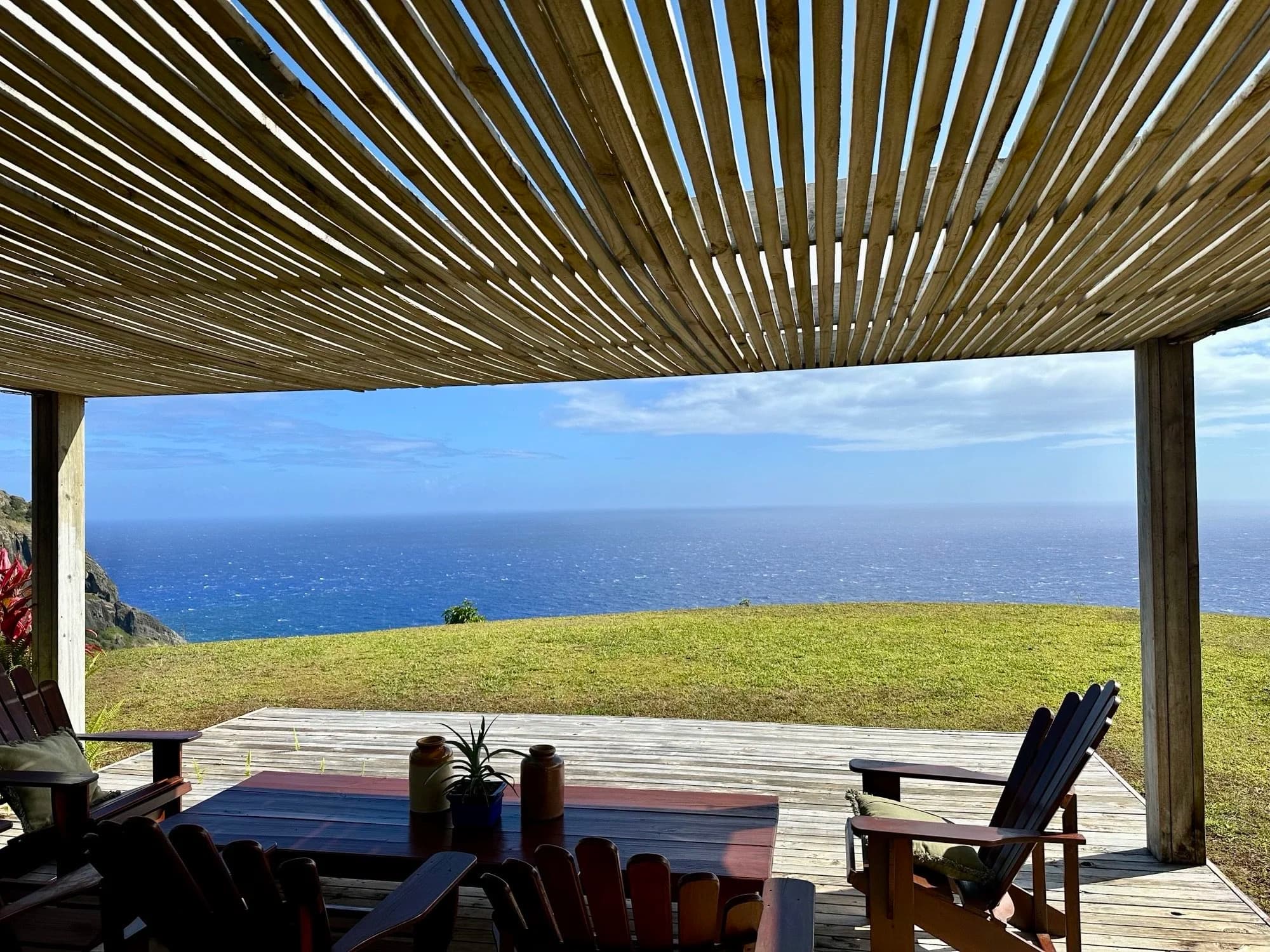 A covered deck with timber chairs looking out over a lawn to the ocean