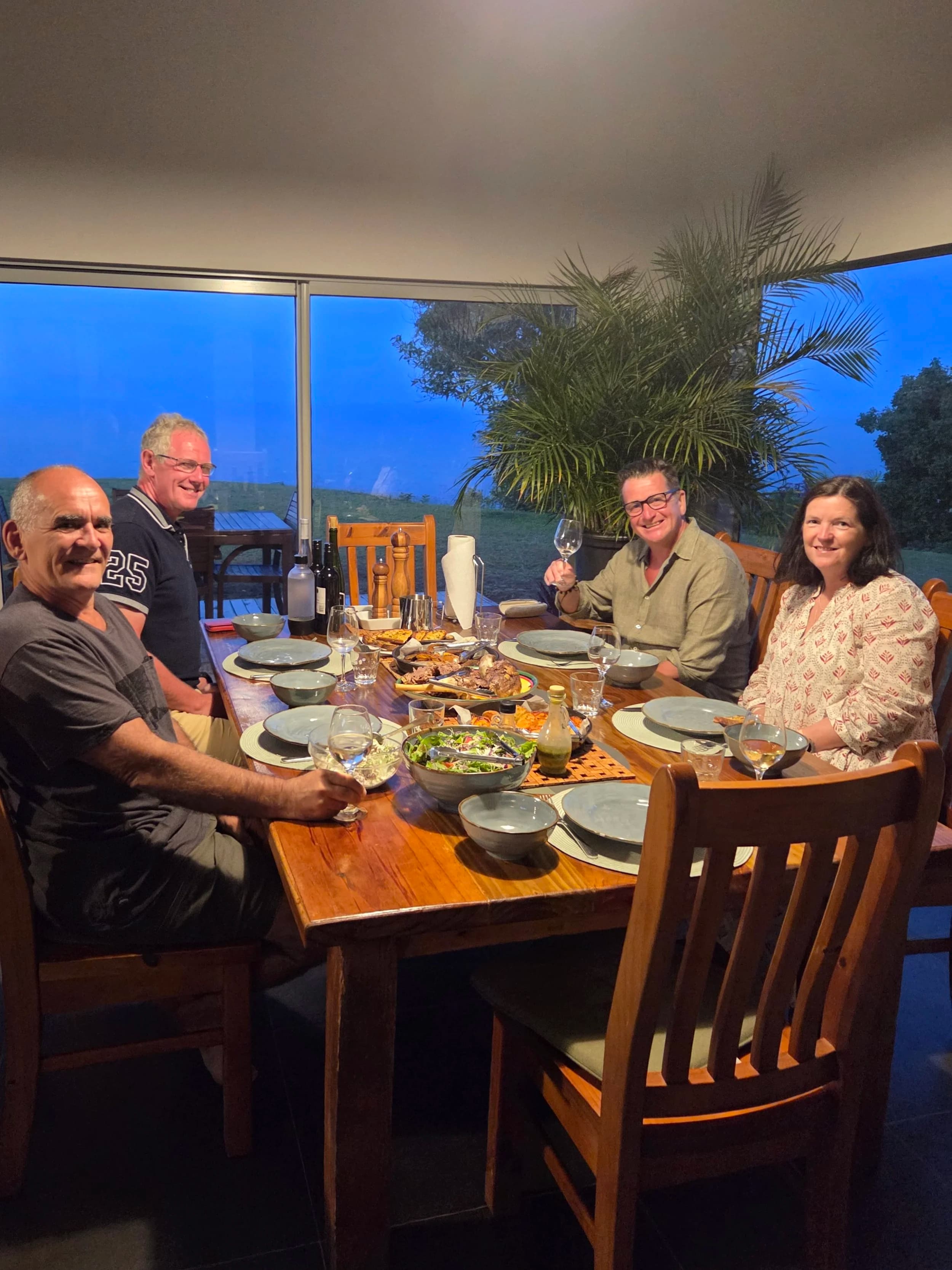 Four people smiling around a dinner table set with food, a window to the garden behind them