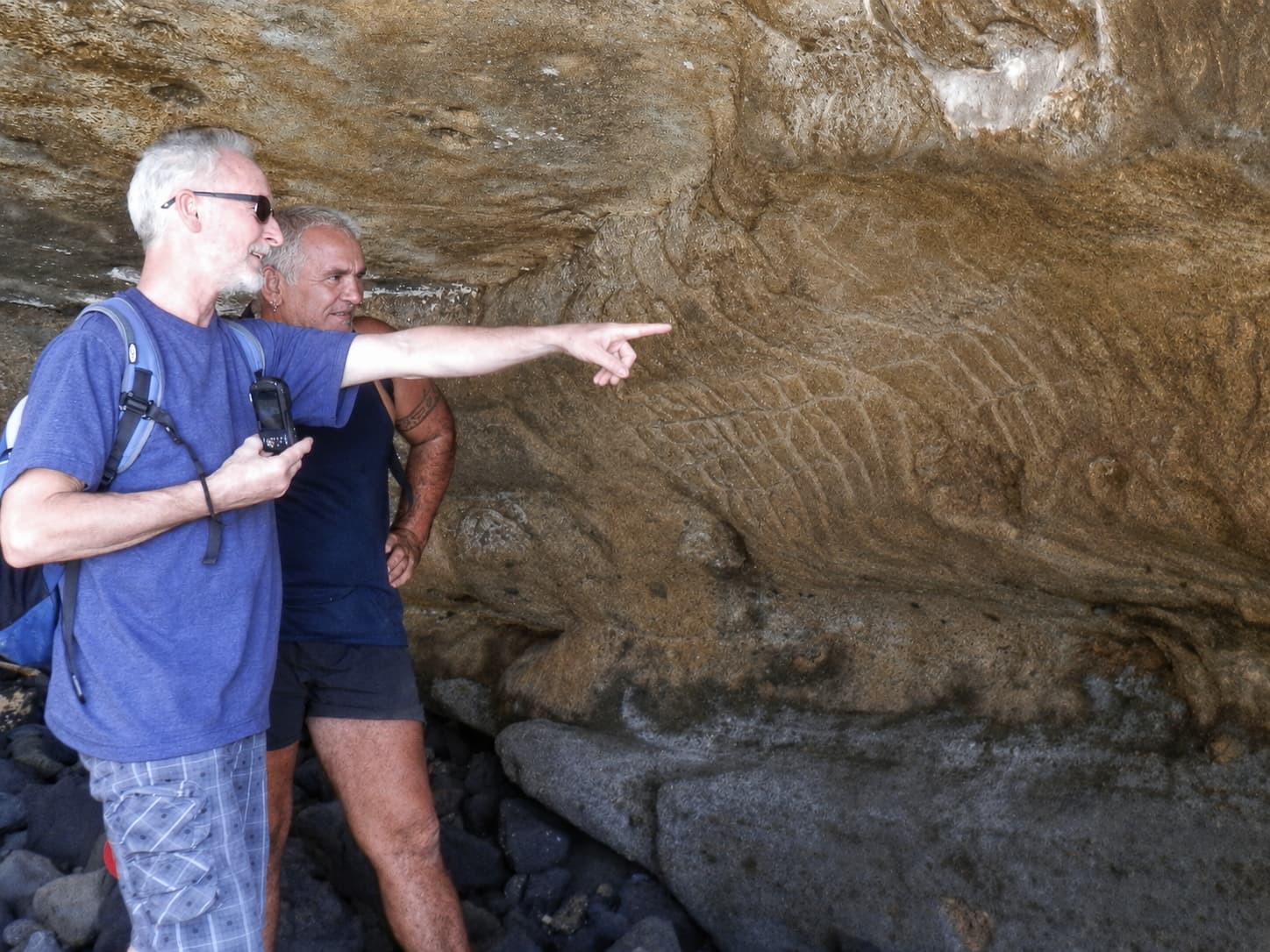 Two men under a rock overhang, one pointing out the petroglyphs carved into it