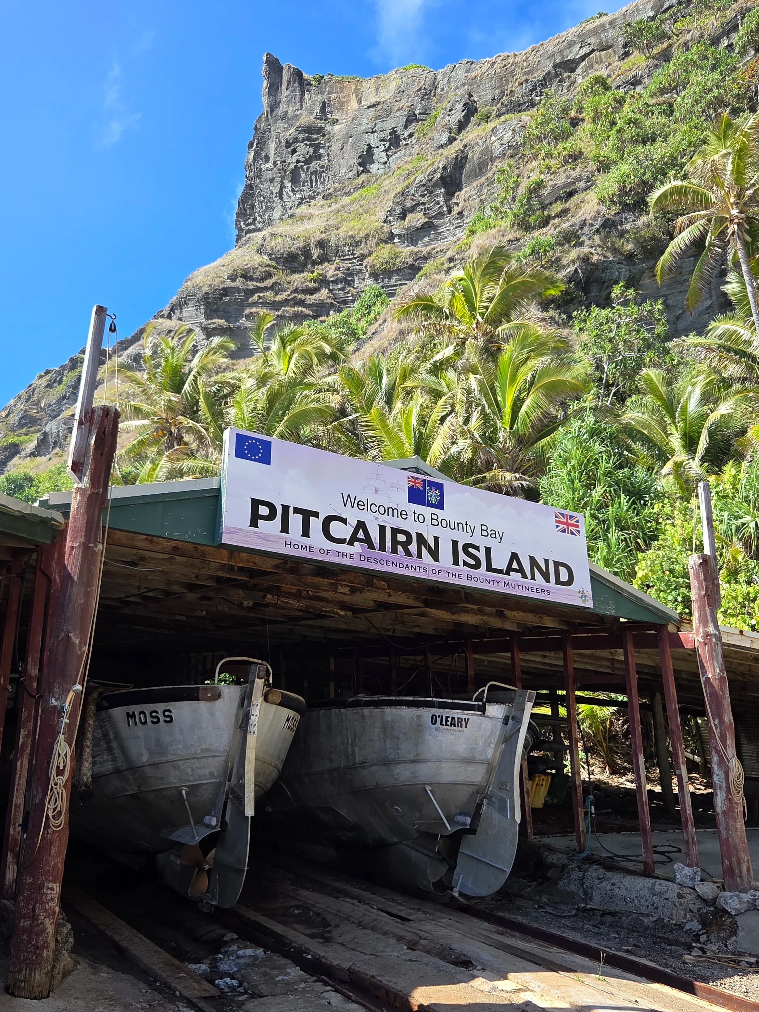 Boats parked under a shelter with a sign that reads 'Welcome to Bounty Bay, Pitcairn Island.' Lush green palm trees and a tall rocky mountain tower in the background.