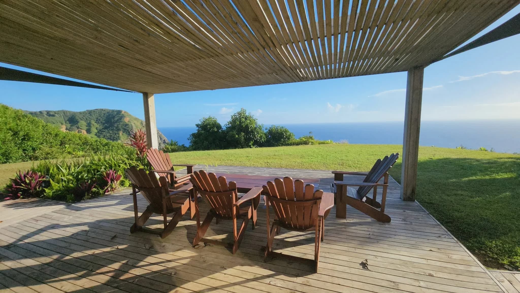 A shaded deck with chairs and a table under a slatted roof, a lawn and the sea beyond