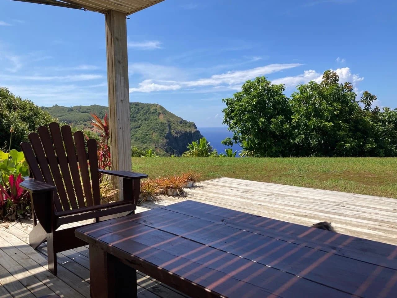 A shaded deck with timber chairs and a table, looking over a lawn to green hills and the sea