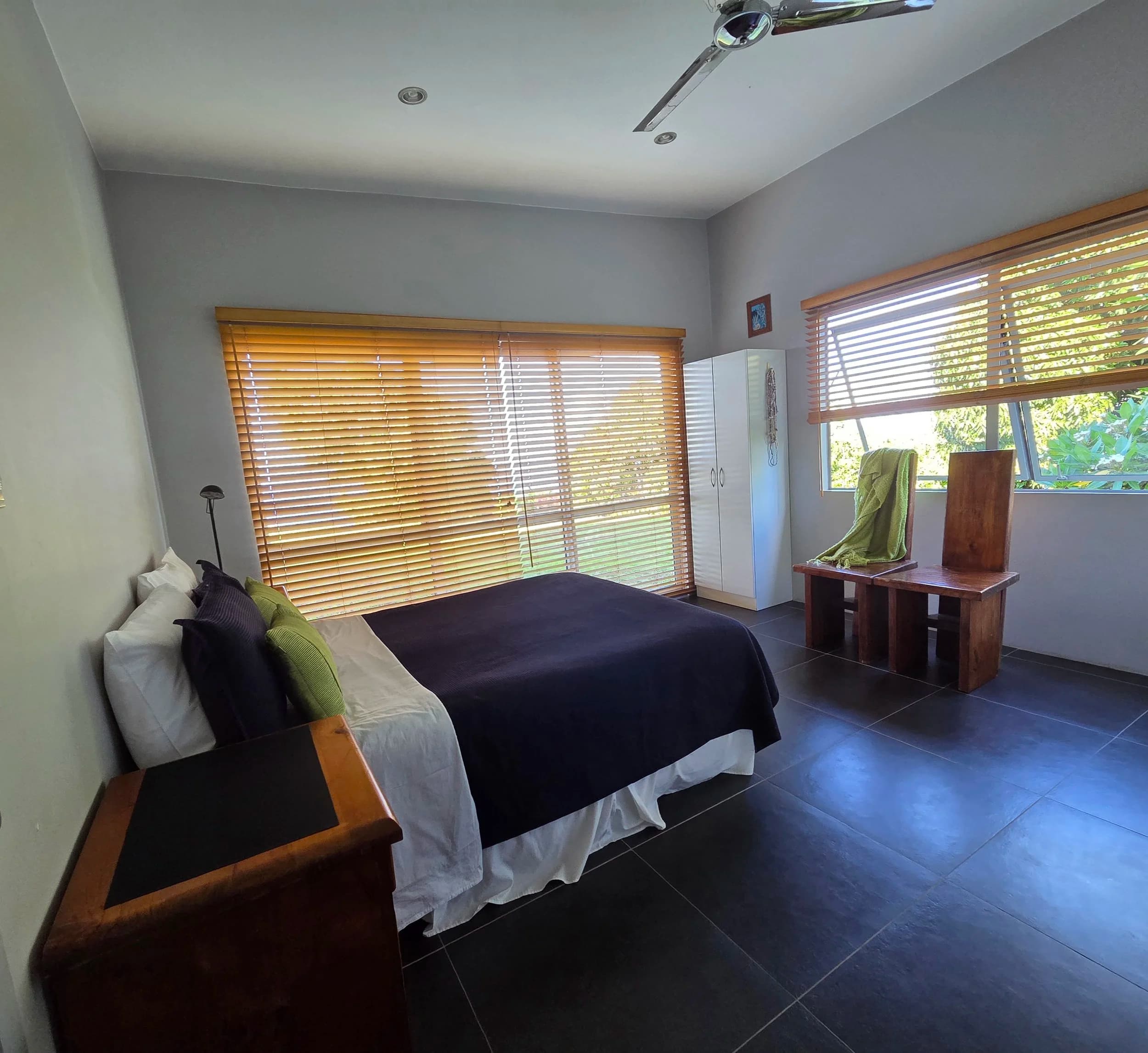 A guest bedroom with a made-up bed, timber blinds and a ceiling fan